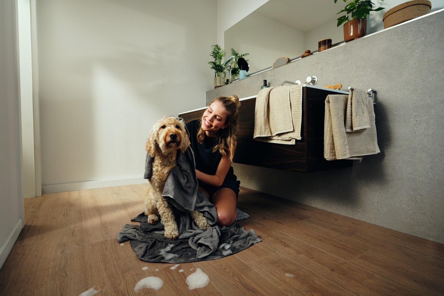 Turin Meşe EL2190 dekorunda EGGER NatureSense Aqua laminat parke üzerinde banyoda köpeğini havluyla kurulayan bir kişi.