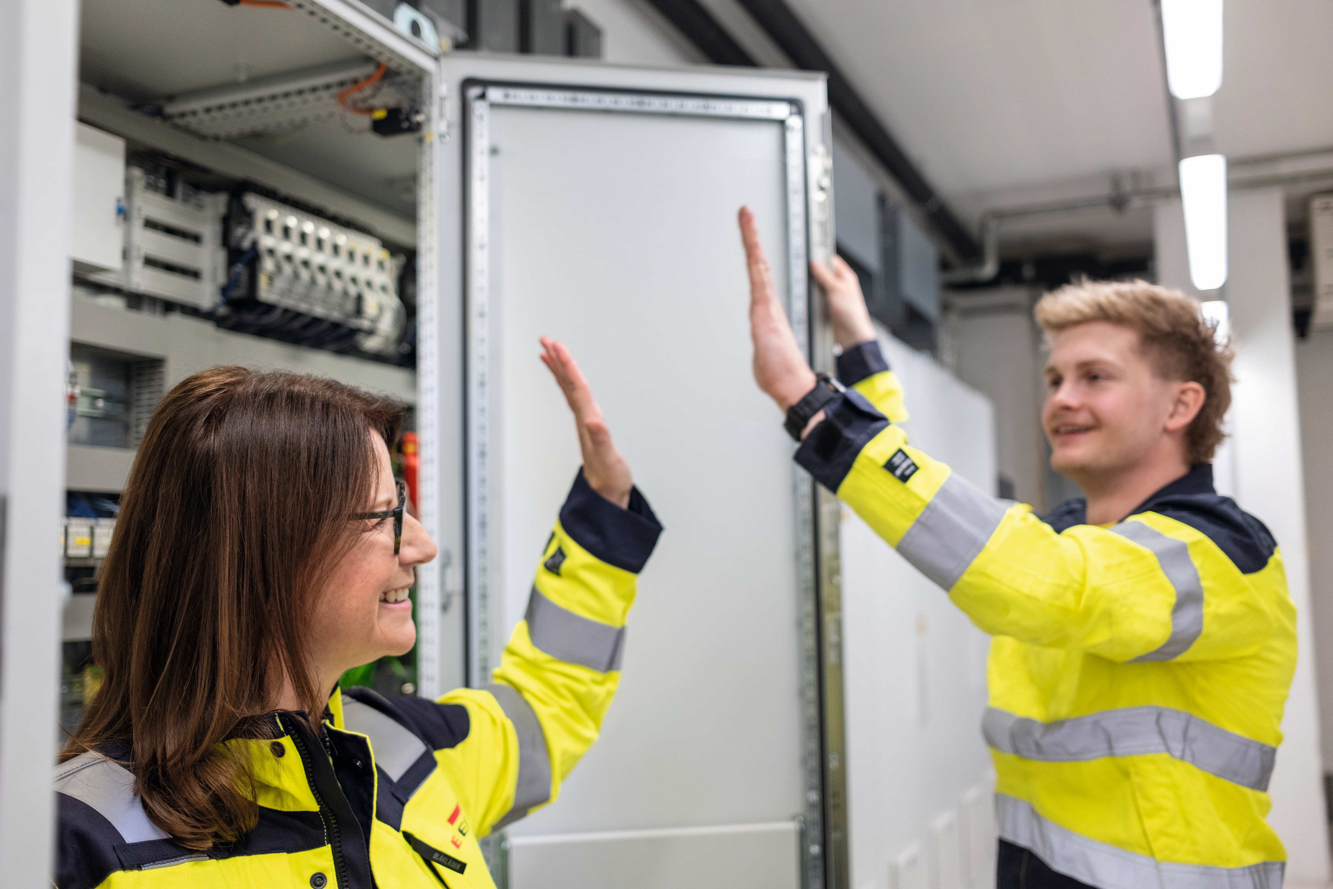 Zwei Personen in gelben Warnjacken heben die Hände für ein High Five vor einem geöffneten Schaltschrank in einem technischen Raum