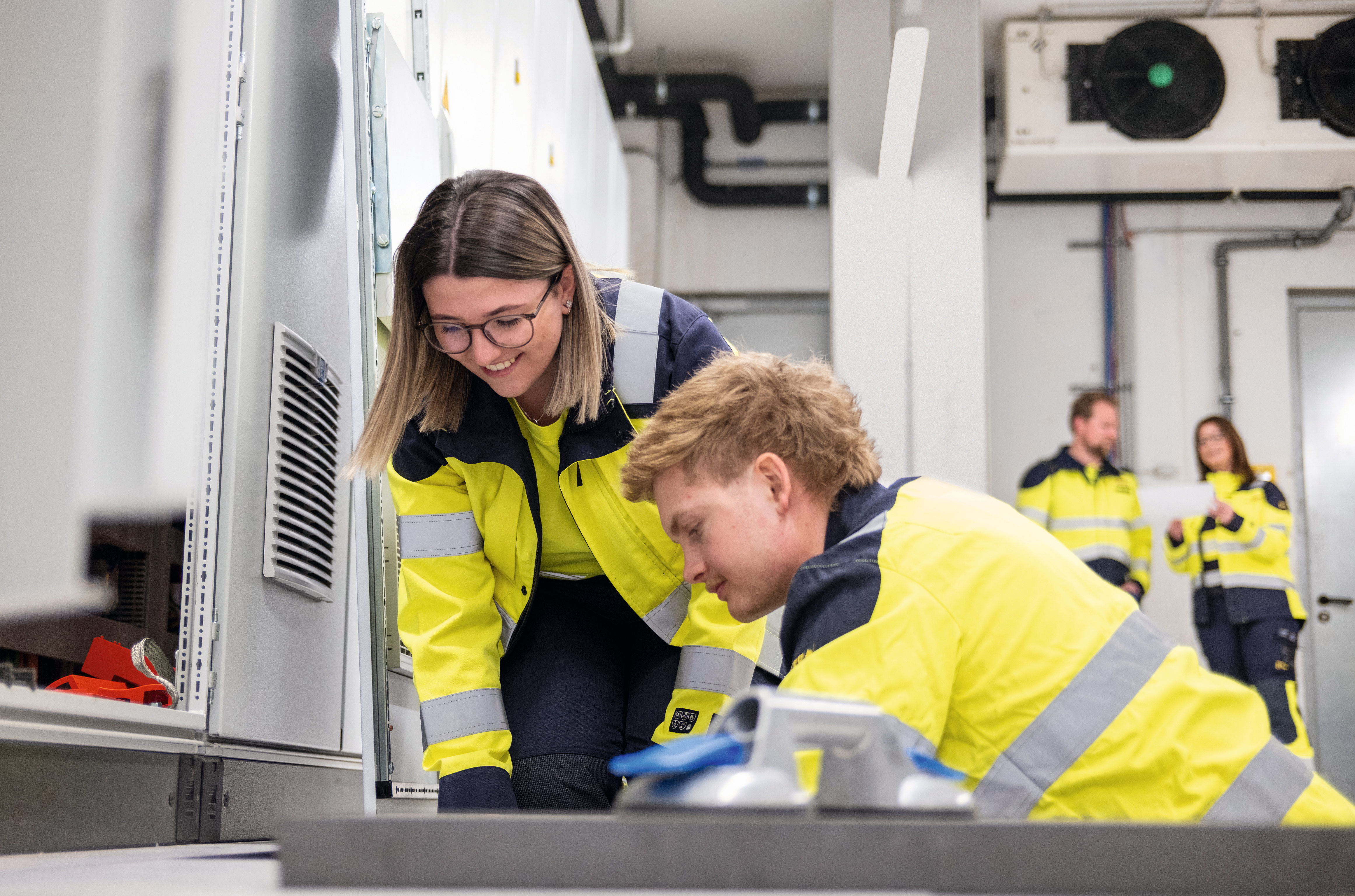 Zwei Personen in gelb-blauen Arbeitsjacken knien vor einem technischen Gerät, im Hintergrund stehen zwei weitere Personen in ähnlicher Arbeitskleidung und unterhalten sich.