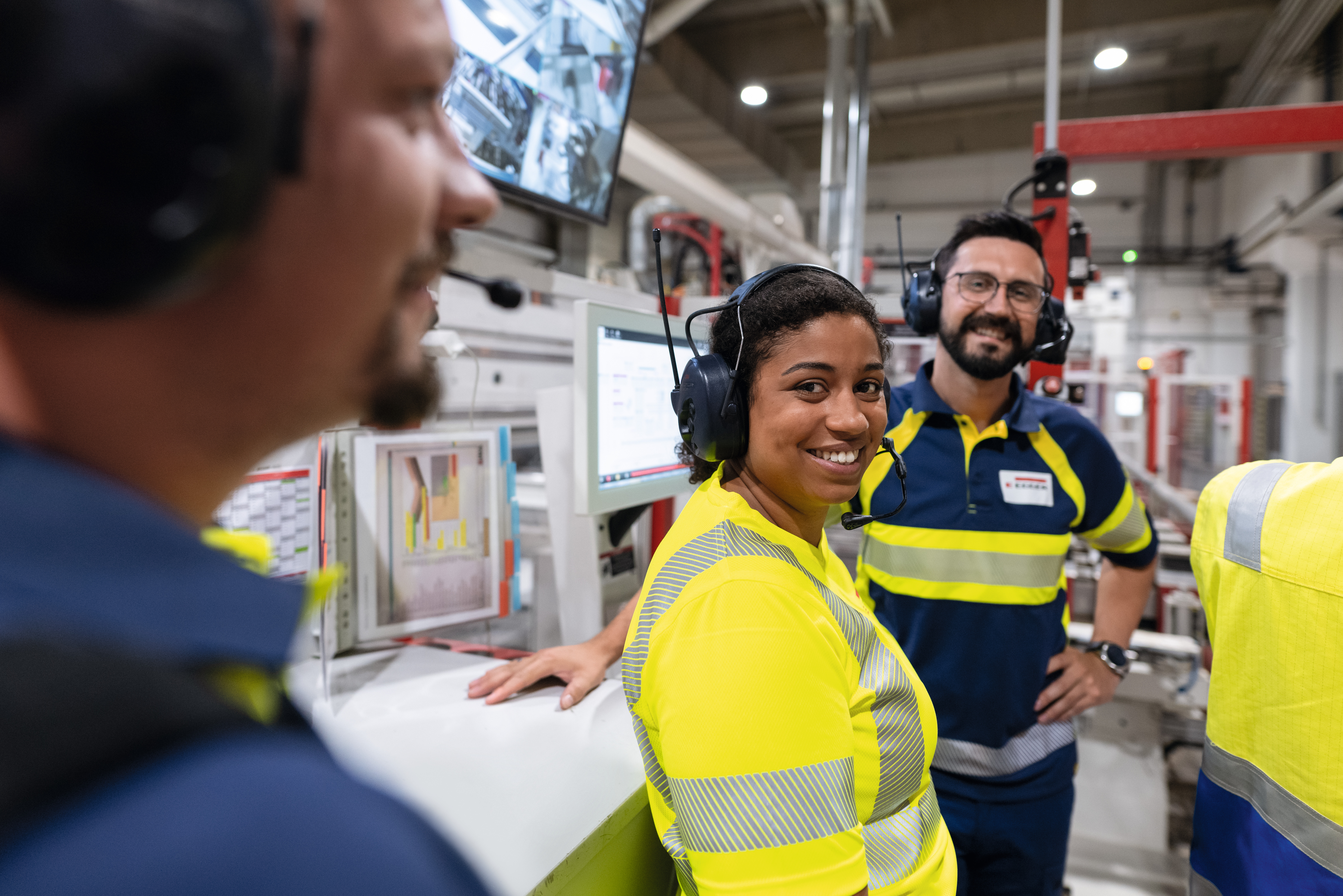 Drei Mitarbeiter:innen in einer Unterhaltung in einem Produktionsbereich Drei Mitarbeiter:innen in einer Unterhaltung in einem Produktionsbereich